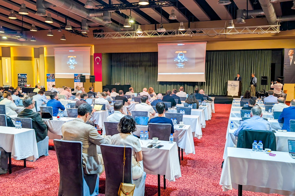 Mehr als 100 Personen sitzen an runden Tischen in einem großen Veranstaltungssaal mit Bühne und Leinwand. Der Saal ist mit roten und weißen Dekorationselementen gestaltet, auf der Bühne stehen Vorhänge und Präsentationstechnik.