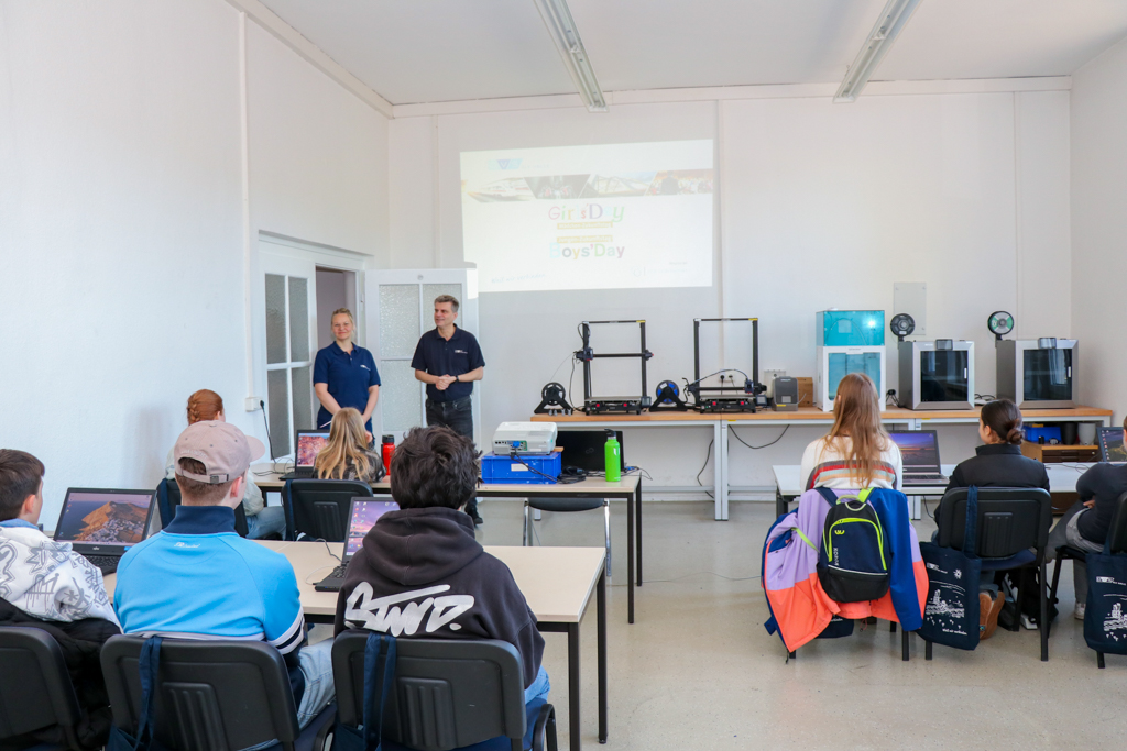 Mehrere Kinder sitzen an Tischen in einem Seminarraum und schauen nach vorne auf eine Präsentationsfläche. Im Raum stehen Schulungstische mit Laptops und Materialien, im Hintergrund sind 3D-Drucker und eine Tafel sichtbar.