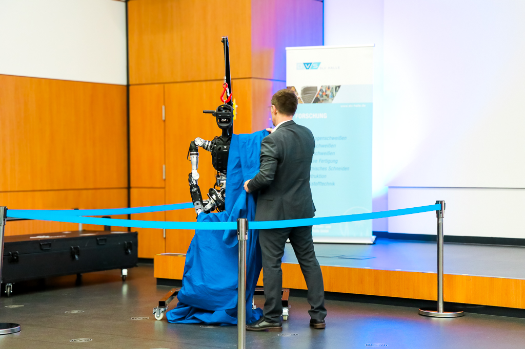 Ein Mann im Anzug entfernt eine blaue Stoffabdeckung von einem humanoiden Roboter auf einer Bühne.
