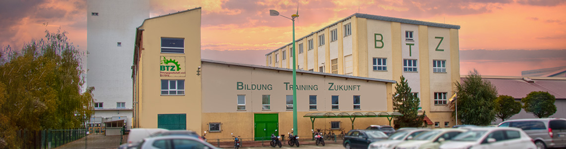 Das Bild zeigt das BTZ-Gebäude mit der Aufschrift „Bildung Training Zukunft“ bei Sonnenuntergang, im Vordergrund sind mehrere parkende Autos zu sehen.