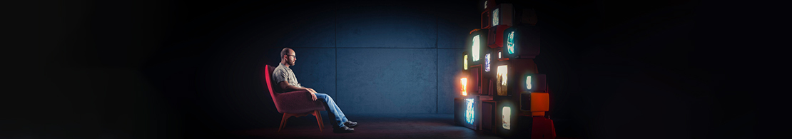 Mann sitzt allein in einem dunklen Raum auf einem Sessel und blickt auf einen hohen Stapel aus alten, leuchtenden Röhrenfernsehern.