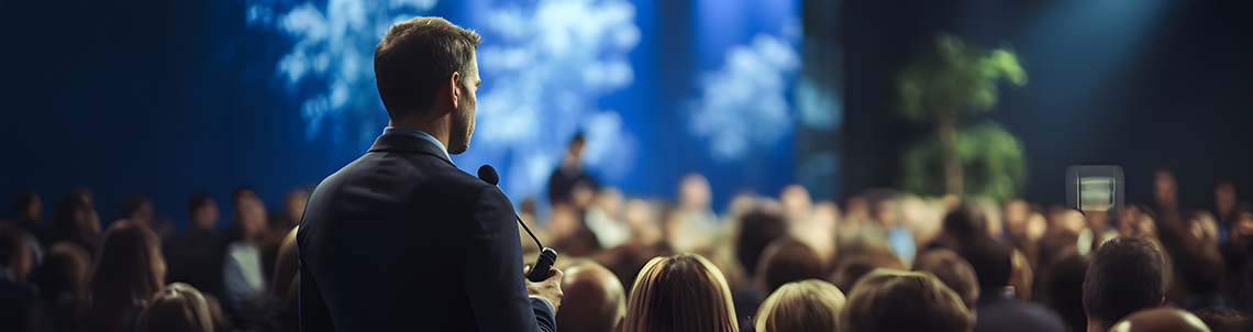 Person spricht vor Publikum auf einer Bühne, im Vordergrund sind zahlreiche sitzende Zuhörende in einem abgedunkelten Raum zu sehen