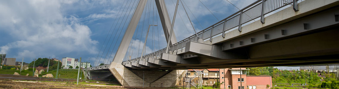 Schrägaufnahme einer modernen Schrägseilbrücke mit massiven Stahl- und Betonelementen, im Vordergrund verlaufen Gleise, im Hintergrund stehen Wohnhäuser und Grünflächen unter wechselhaftem Himmel.