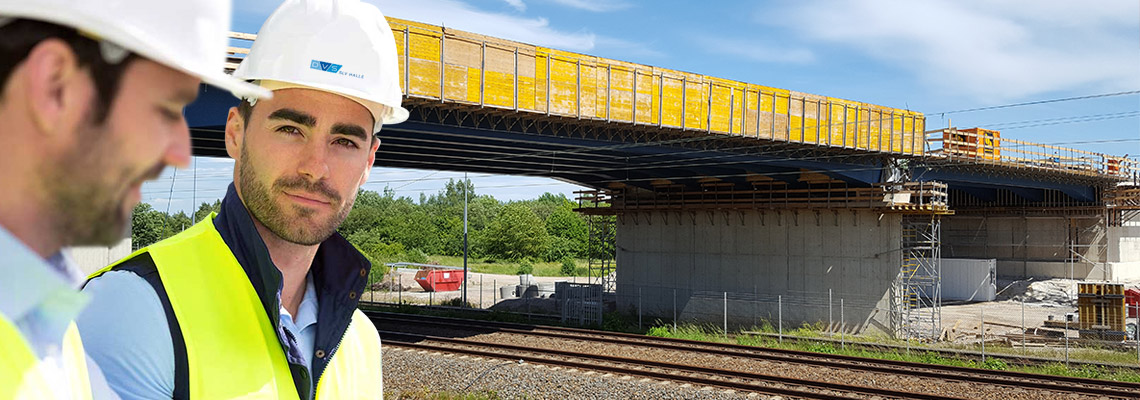 Zwei Männer mit Schutzhelmen und Warnwesten stehen an einer Baustelle vor einer im Bau befindlichen Brücke, im Hintergrund sind Gleise, Gerüste und Betonpfeiler zu sehen.