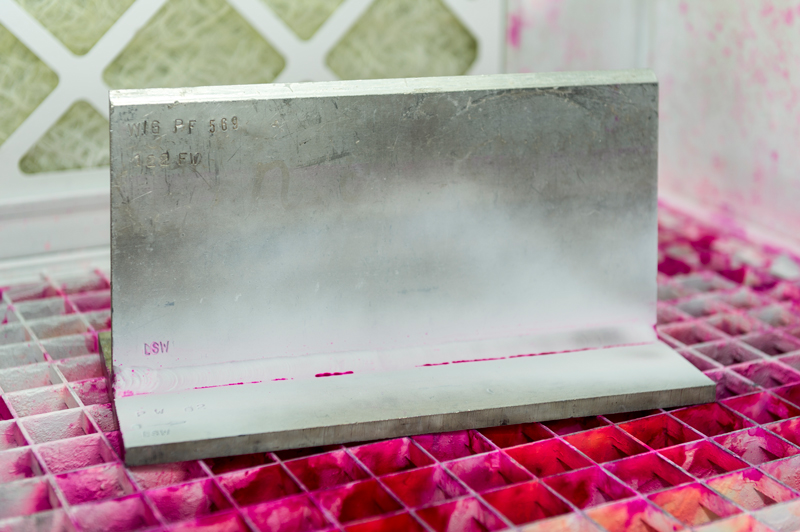 Metallbauteil steht auf einem Gitterrost, darauf sind deutliche pinke Farbanzeigen aus einer Eindringprüfung sichtbar, Farbe hat sich auf der Fläche verteilt.