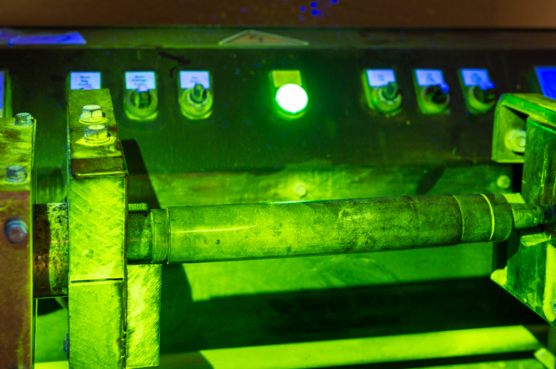Metallzylinder liegt in einer Prüfhalterung und leuchtet unter UV-Licht grünlich, feine Anzeigen der Magnetpulverprüfung sind auf der Oberfläche erkennbar.